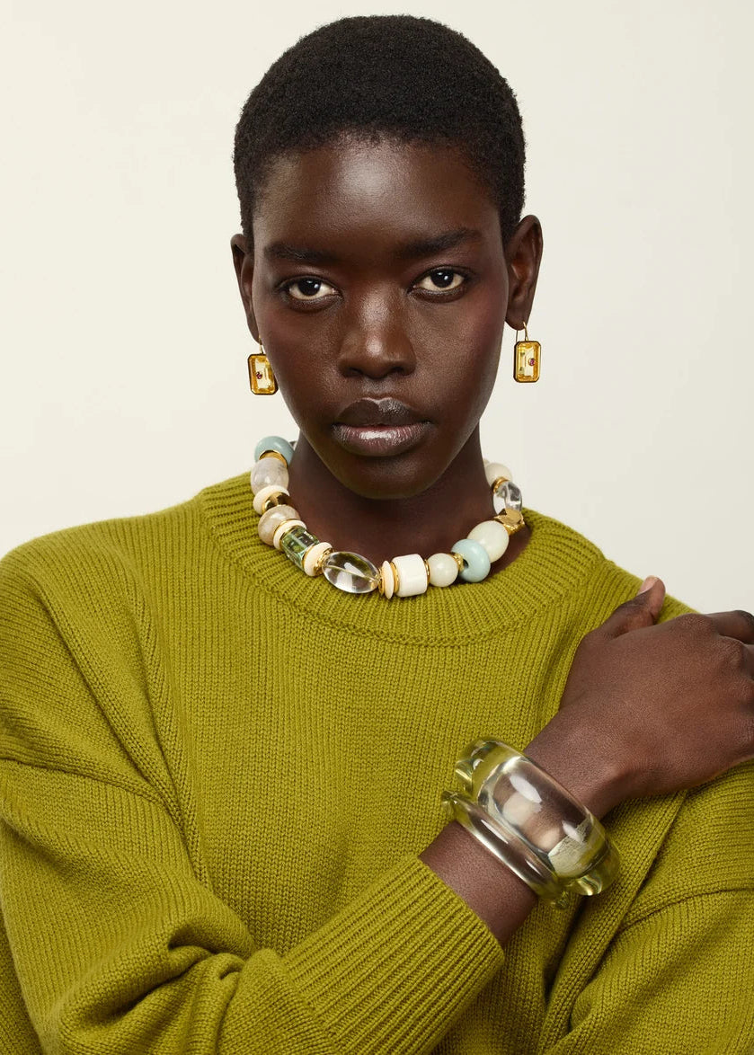 A model with short dark hair is wearing a textured olive green knit sweater, a chunky beaded necklace, gold rectangular earrings, and a wide clear resin cuff bracelet, posed against a solid off-white background.