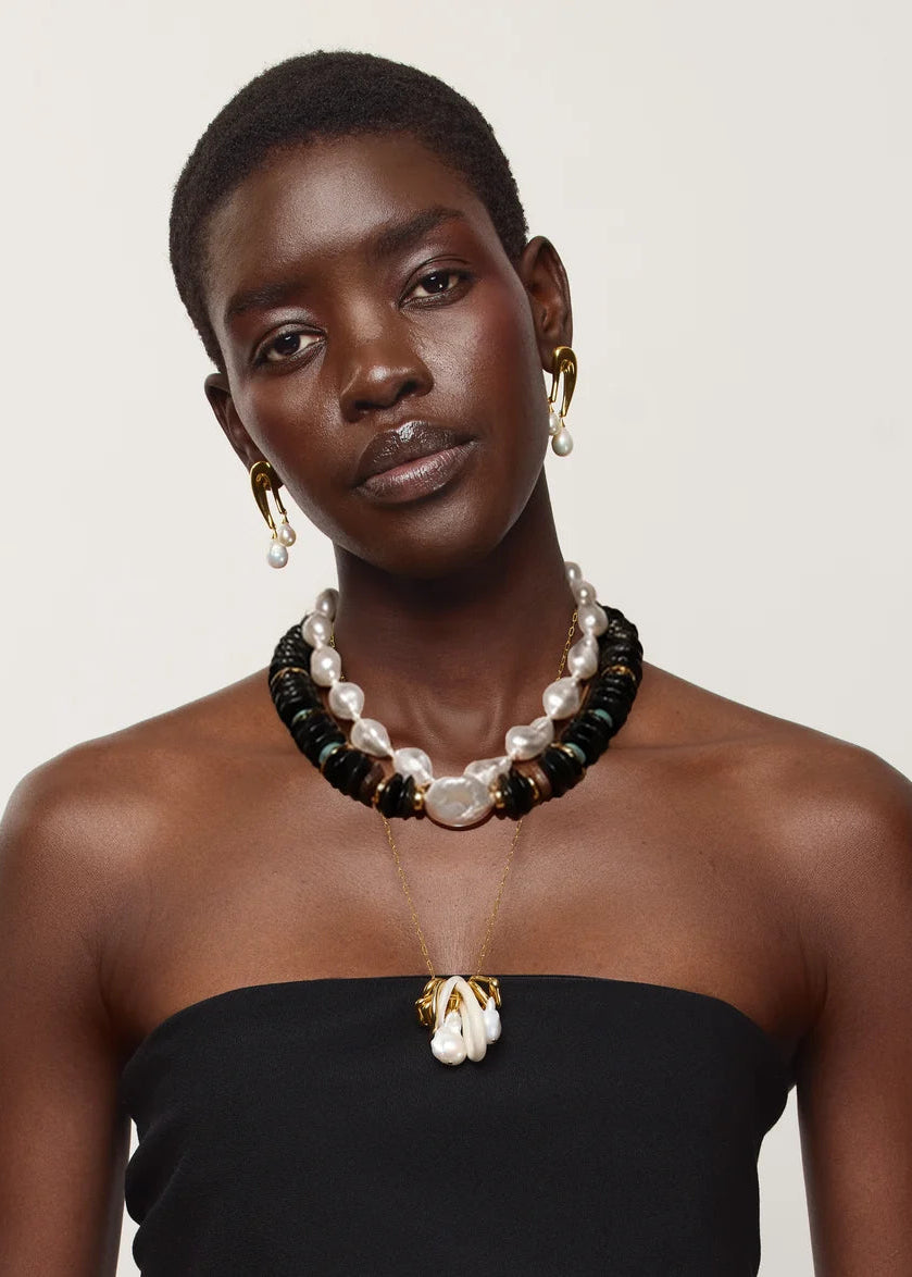 A model wearing a multi-strand necklace with white pearls, black beads, and gold-toned accents, along with matching gold hoop earrings and a gold pendant necklace, styled with a black strapless top against a light beige background.
