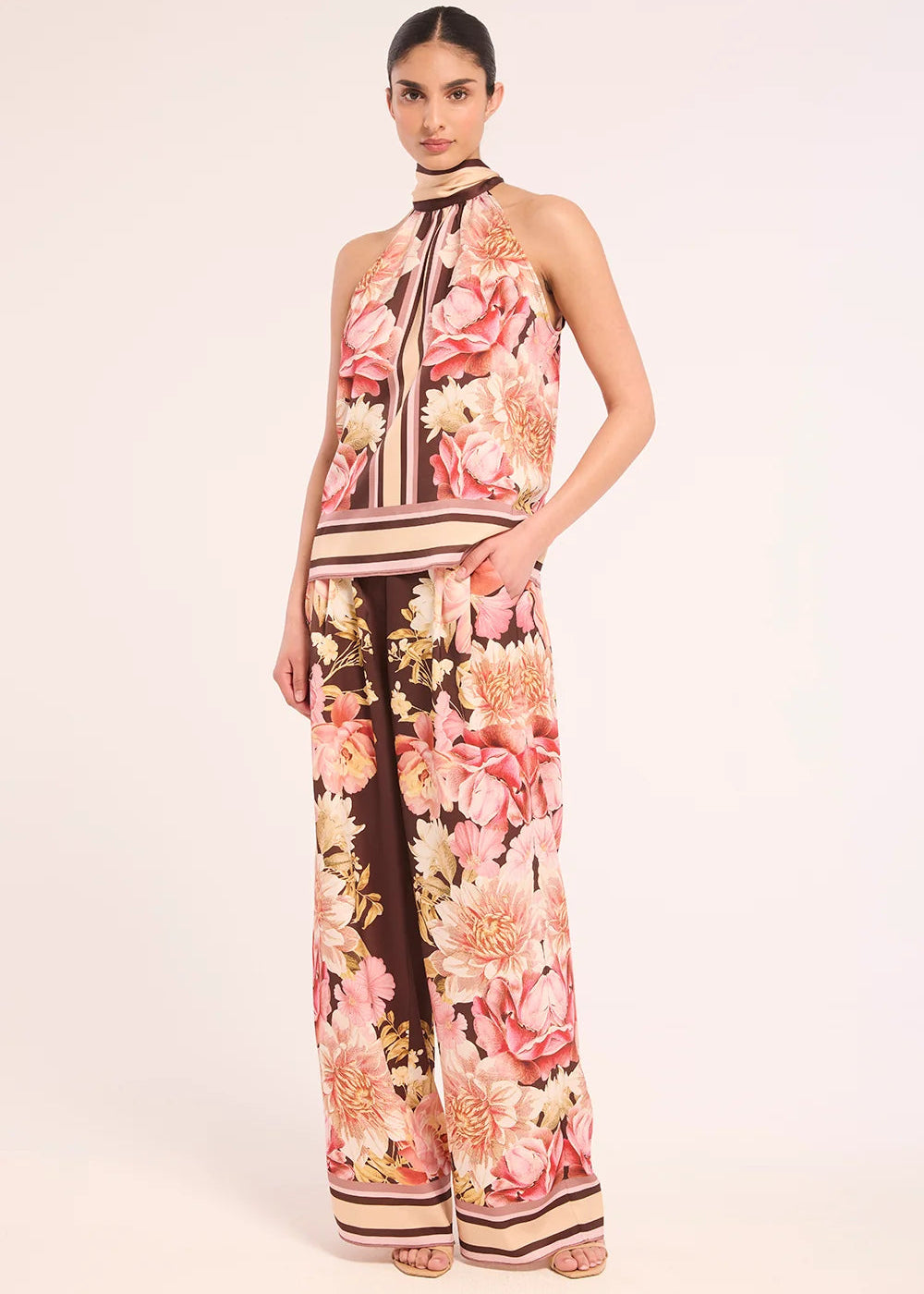 A model wearing a sleeveless halter-neck jumpsuit with a pink, peach, and cream floral print over a dark brown base, styled with neutral striped trim, set against a plain light beige studio background.