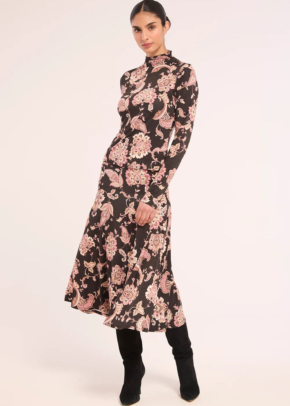 A woman models a long-sleeved midi dress with a black base and a pink floral paisley pattern, paired with black boots, against a plain light background.