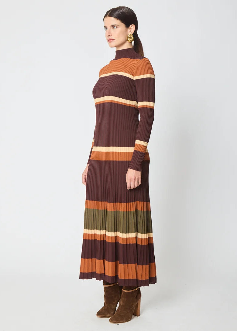 A woman modeling a long-sleeve ribbed knit midi dress with horizontal stripes in dark brown, rust orange, olive green, and cream, paired with brown suede ankle boots against a light gray studio background.