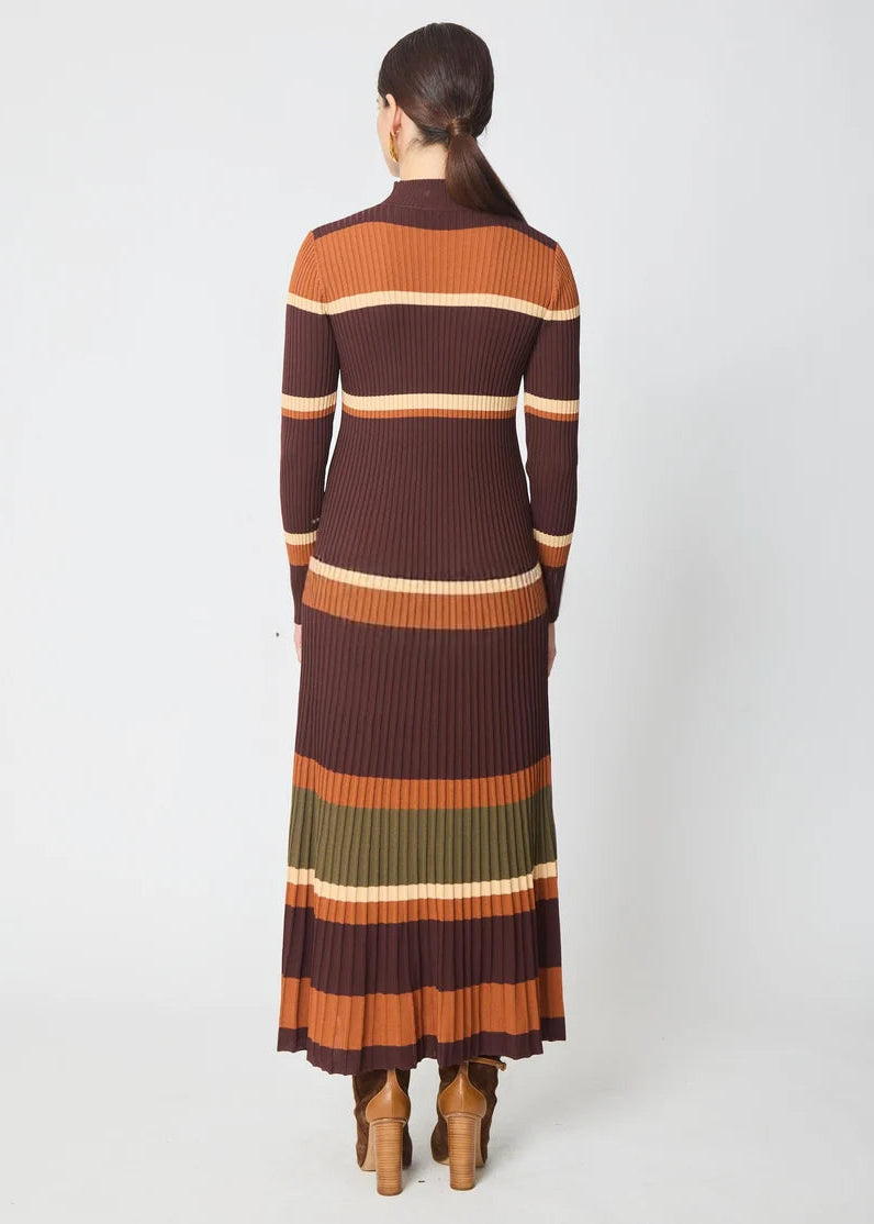 A woman viewed from the back wearing a long-sleeve ribbed knit midi dress with horizontal stripes in dark brown, rust orange, olive green, and cream, paired with brown heeled boots against a light gray studio background.