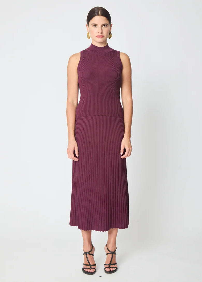 A woman modeling a sleeveless ribbed knit midi dress in a deep burgundy/plum color, paired with black strappy high-heeled sandals, against a plain light gray studio background.