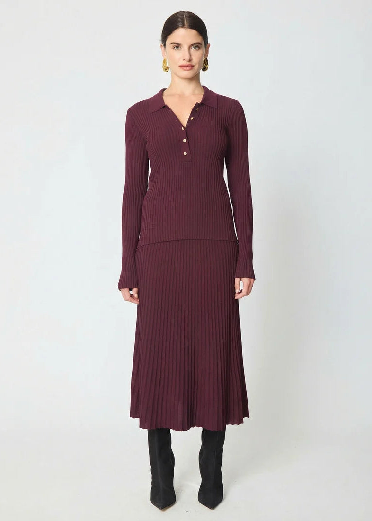 A woman modeling a ribbed knit long-sleeve polo-neck midi dress in deep burgundy, paired with black ankle boots, against a plain light gray studio background.