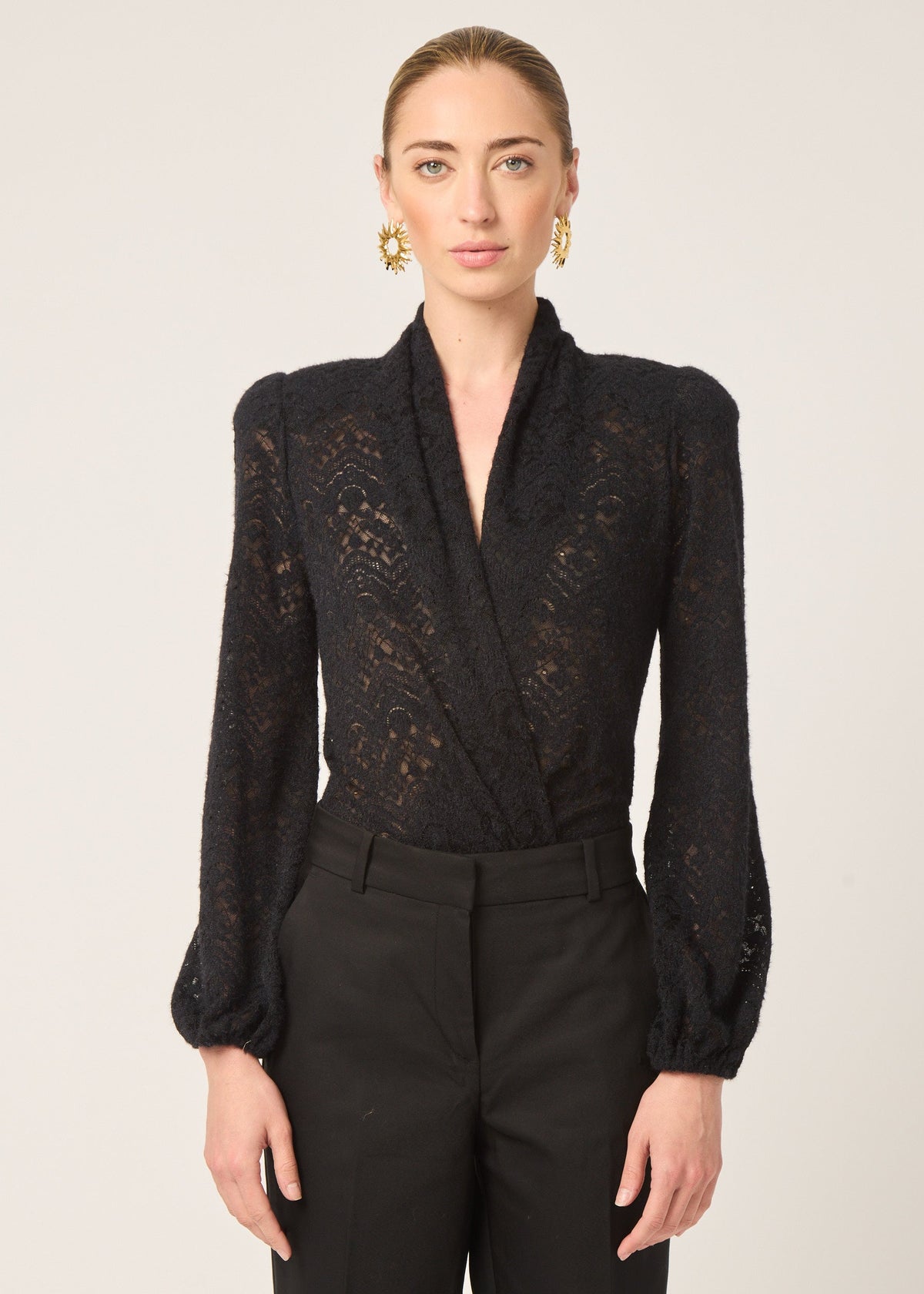 A woman modeling a black long-sleeve lace wrap top with structured shoulders and black trousers against a plain light background.