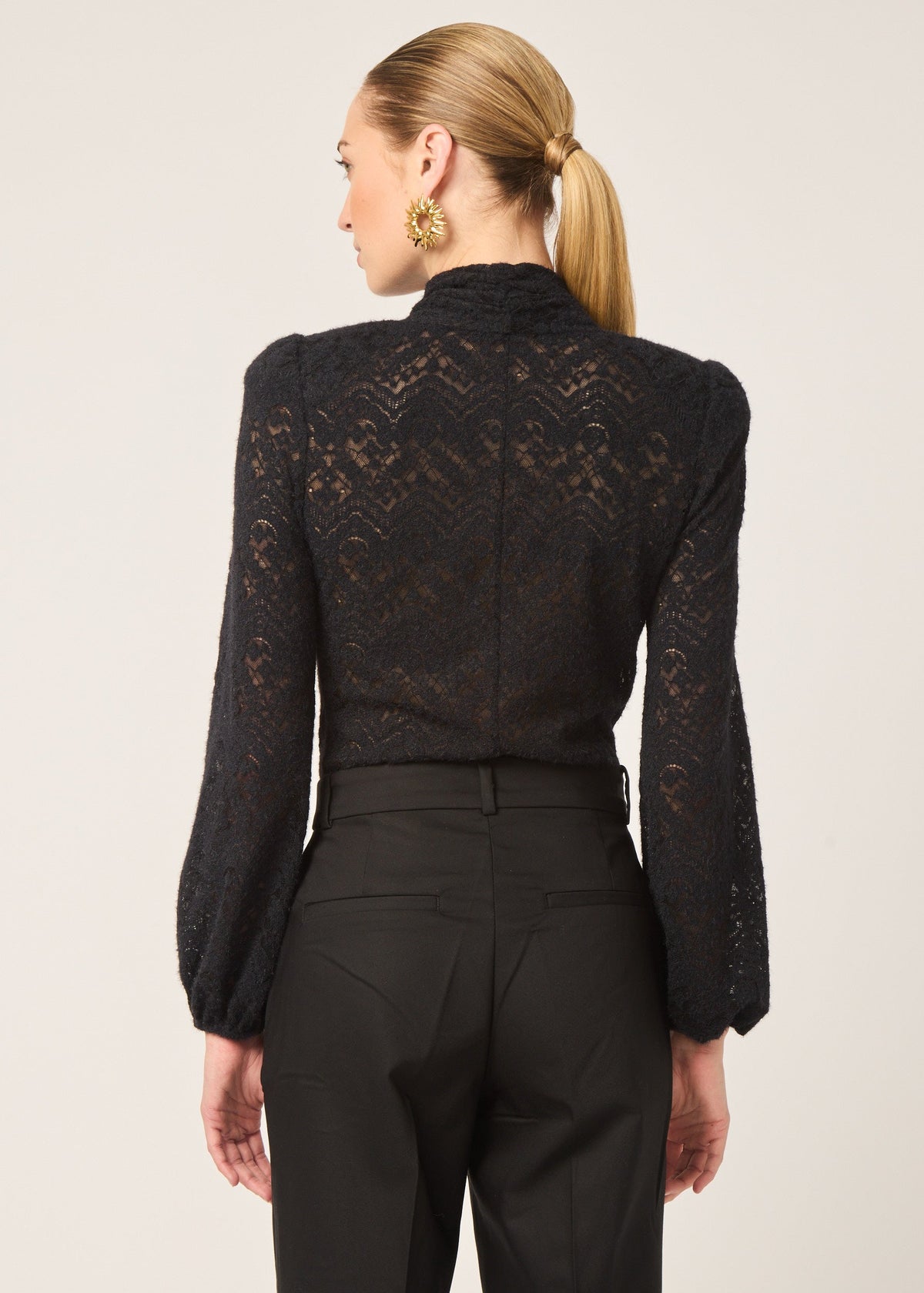 A woman shown from the back wearing a black lace long-sleeve mock-neck top and black trousers, against a plain light beige studio background.