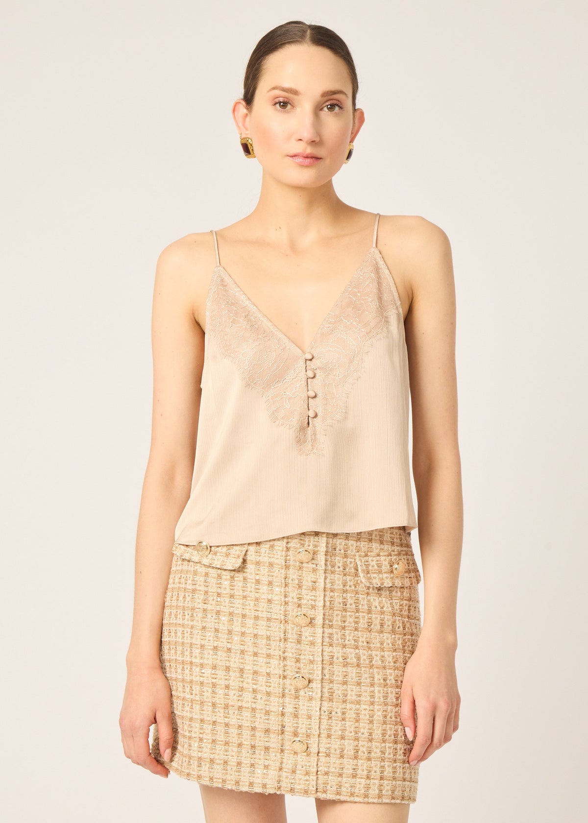 A model wearing a beige lace-trimmed camisole top and a beige and brown tweed mini skirt, photographed against a solid light beige studio background.