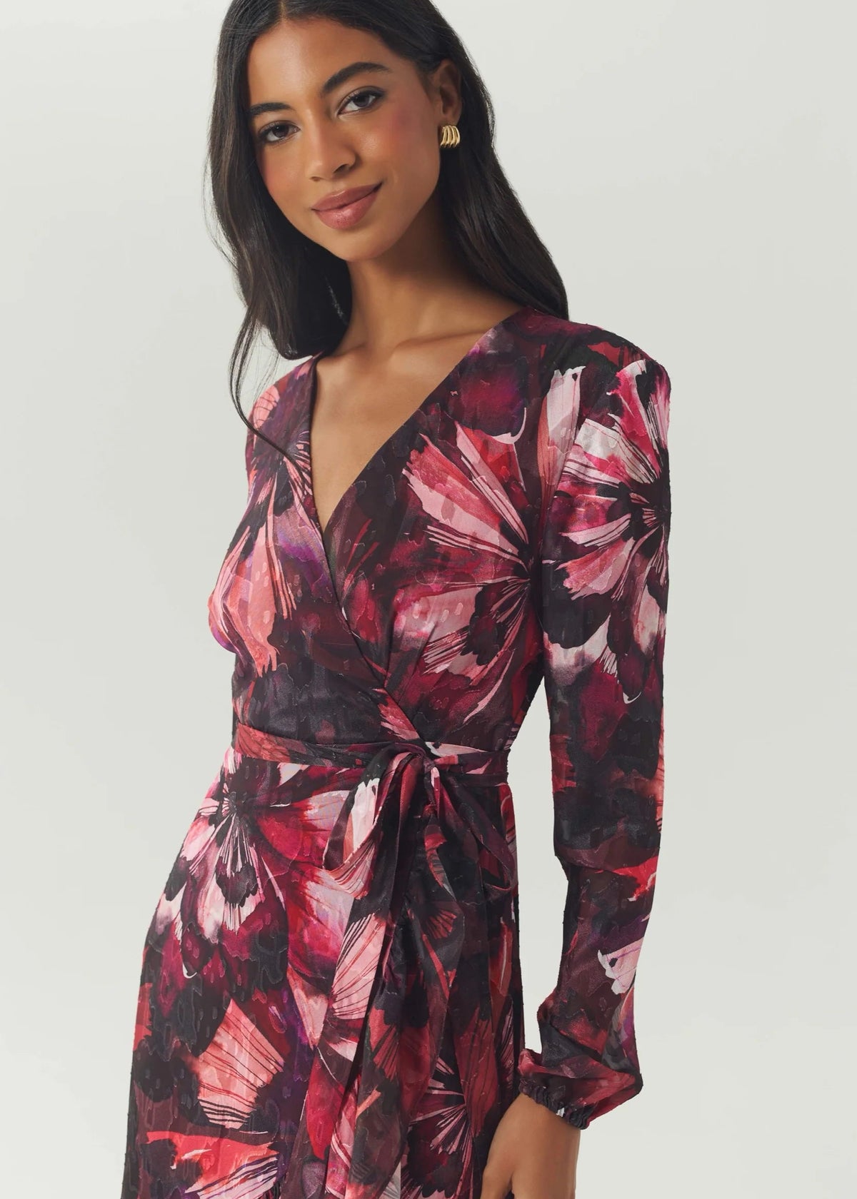 A woman wearing a long-sleeve wrap dress with a dark background and a pink, red, and purple floral print, photographed against a light gray studio background.