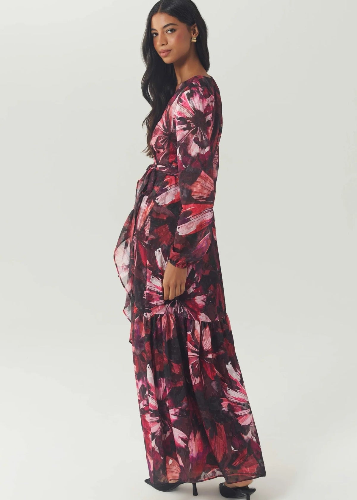 A woman modeling a long-sleeved maxi dress with a dark pink, red, and black floral print, shown from a side-back angle against a light gray studio background.
