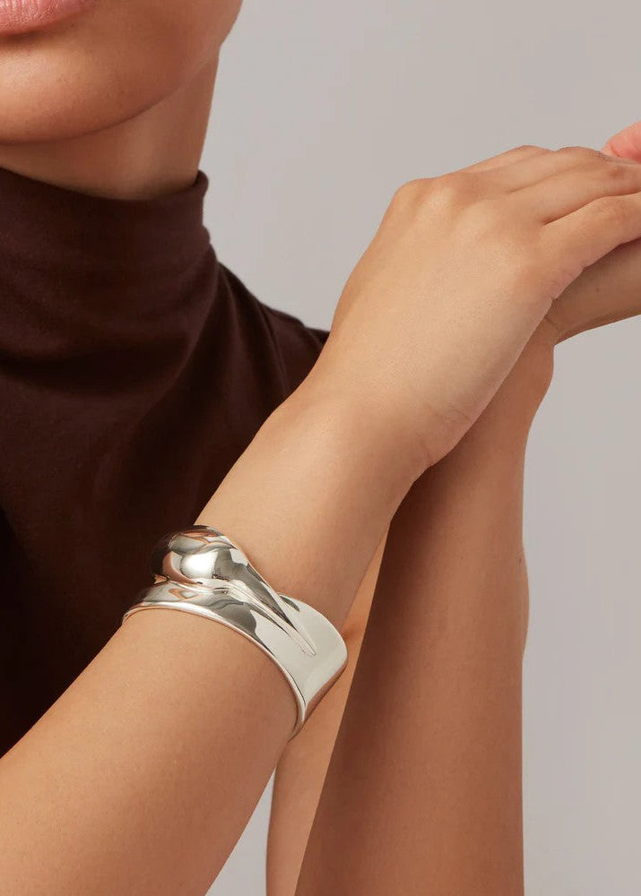A close-up of a model's wrist wearing a wide, polished silver cuff bracelet, paired with a dark brown turtleneck top against a neutral light gray background.