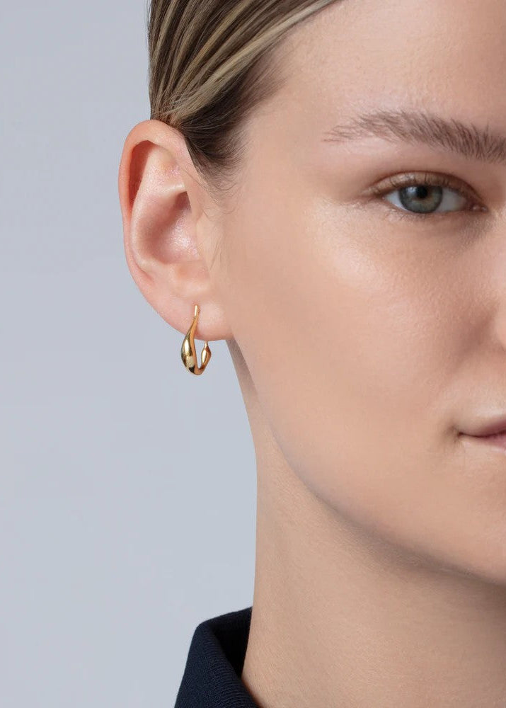 A close-up of a model's ear wearing a small gold hoop earring against a light gray background.