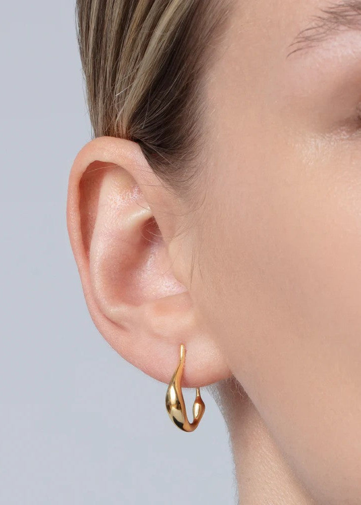 A close-up of a model's ear wearing a small, polished gold huggie hoop earring against a light gray background.