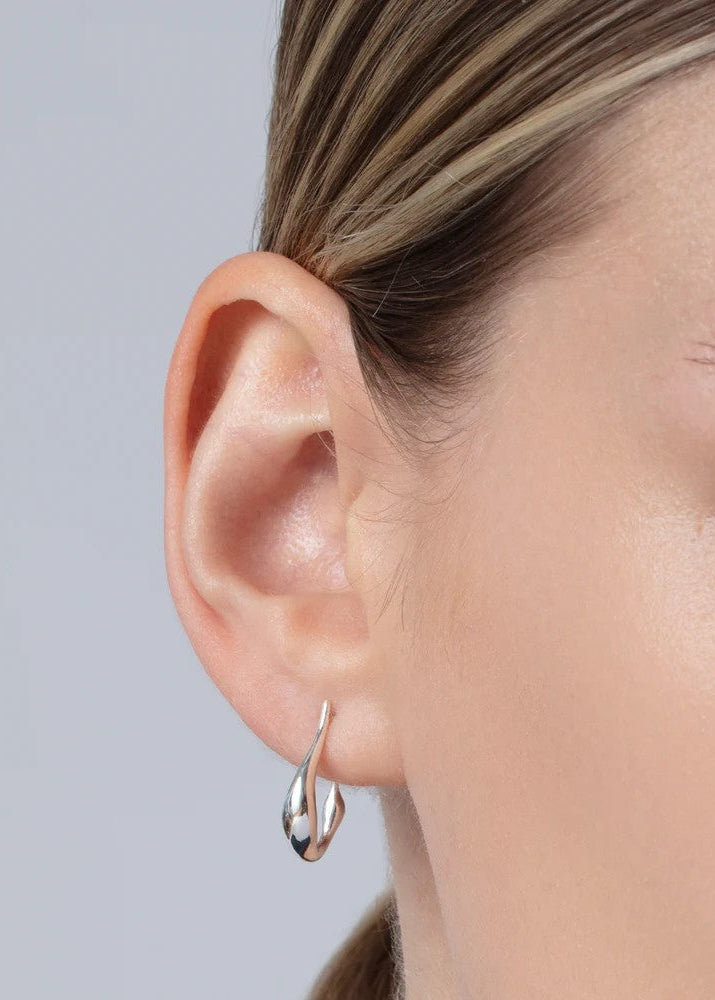 A close-up of a model's ear wearing a small, polished silver hoop earring with a curved, teardrop-like design.