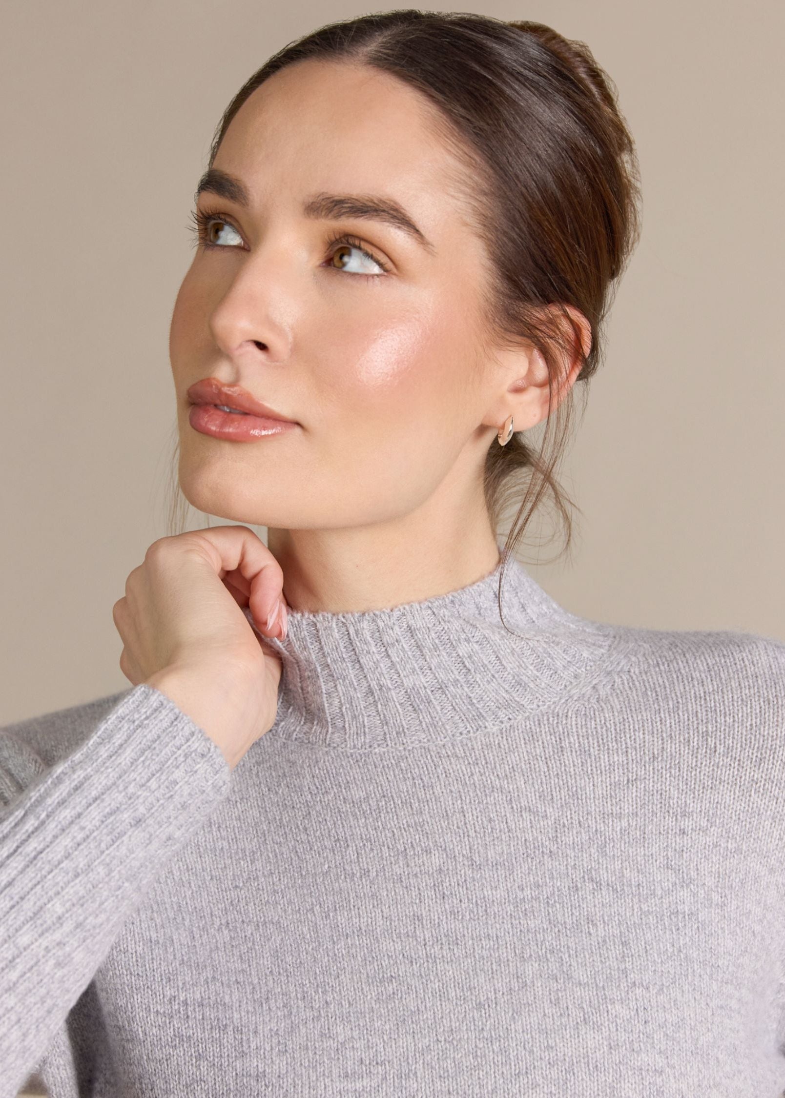 A woman wearing a light gray ribbed knit mock neck sweater, shown in a close-up portrait against a solid beige background.