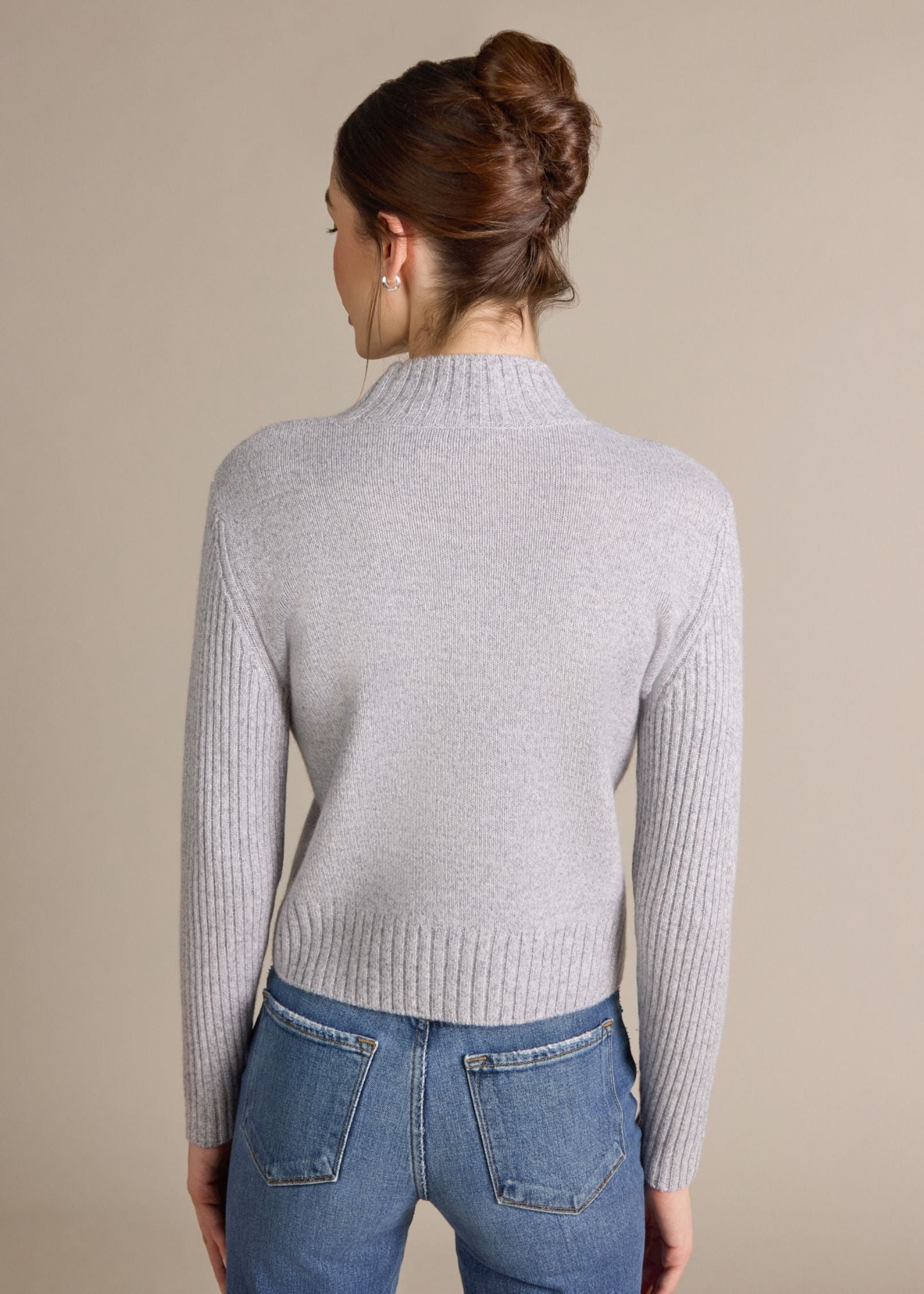 A woman shown from the back wearing a light gray ribbed mock neck sweater and blue jeans, set against a plain beige studio background.