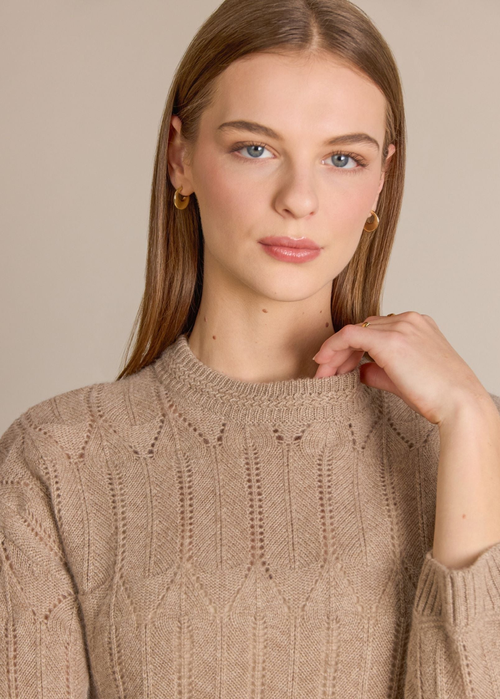 A woman wearing a beige cable-knit crewneck sweater, shown from the chest up against a plain beige studio background.