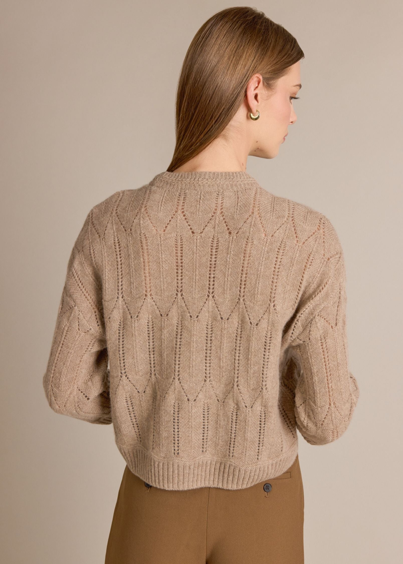 A woman shown from the back wearing a beige cable-knit cropped sweater and brown pants against a plain beige studio background.