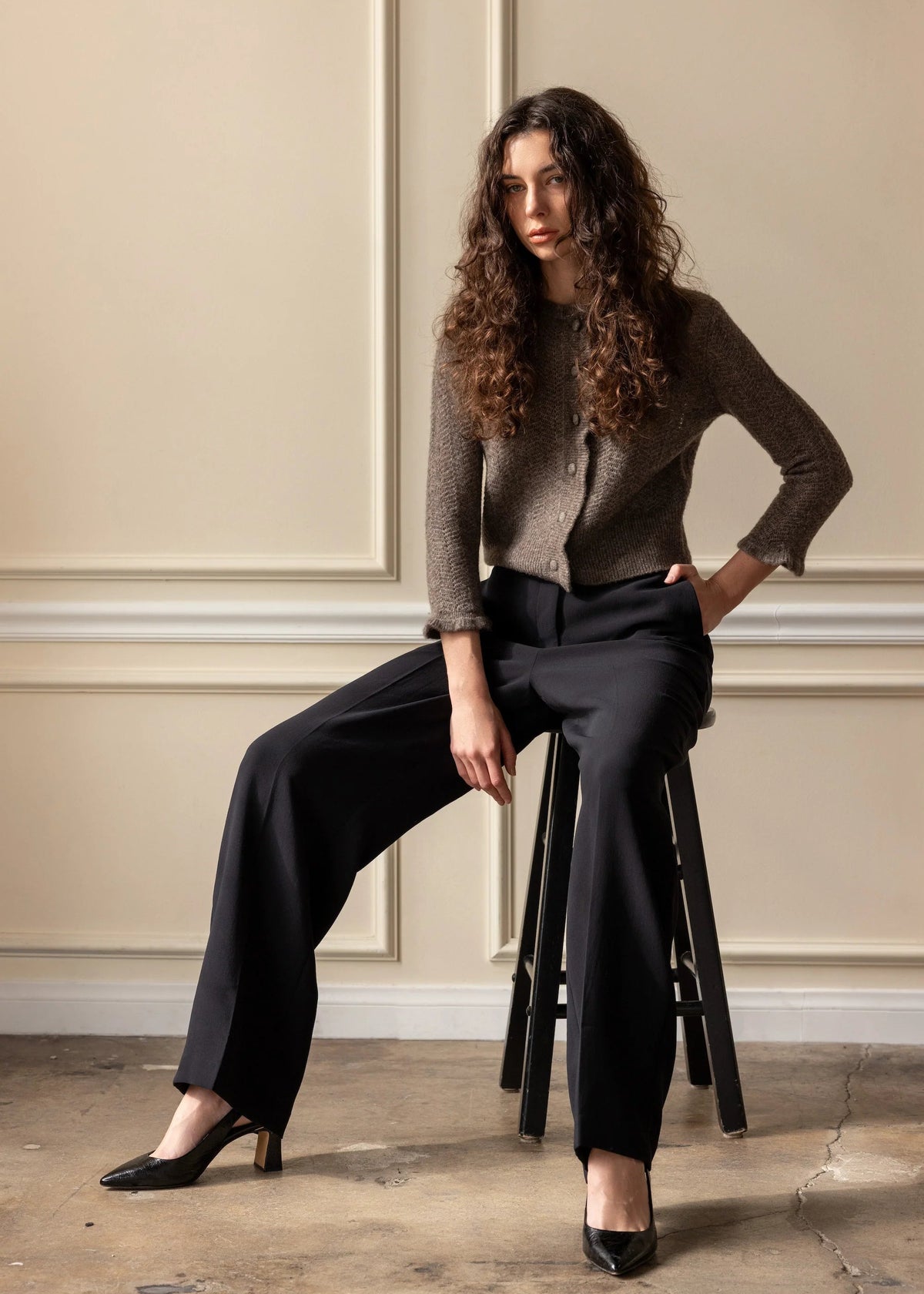 A woman with long curly brown hair sits on a black stool in a room with beige paneled walls and a concrete floor. She is wearing a brown textured knit cardigan, black wide-leg trousers, and black pointed-toe heels.