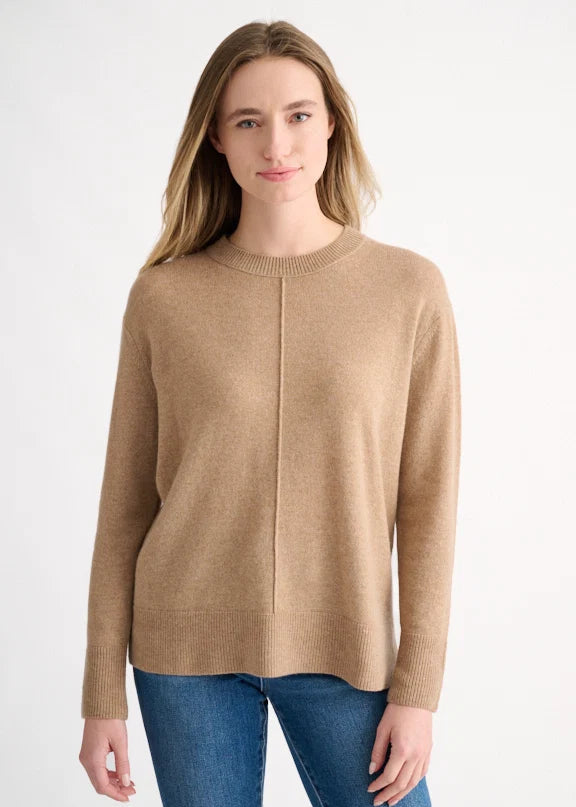 A woman wearing a light tan crewneck knit sweater and blue jeans, standing against a plain white studio background.