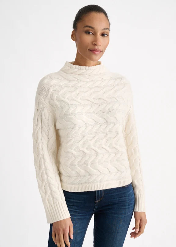 A woman wearing a cream cable-knit mock neck sweater and dark blue jeans, standing against a plain white studio background.