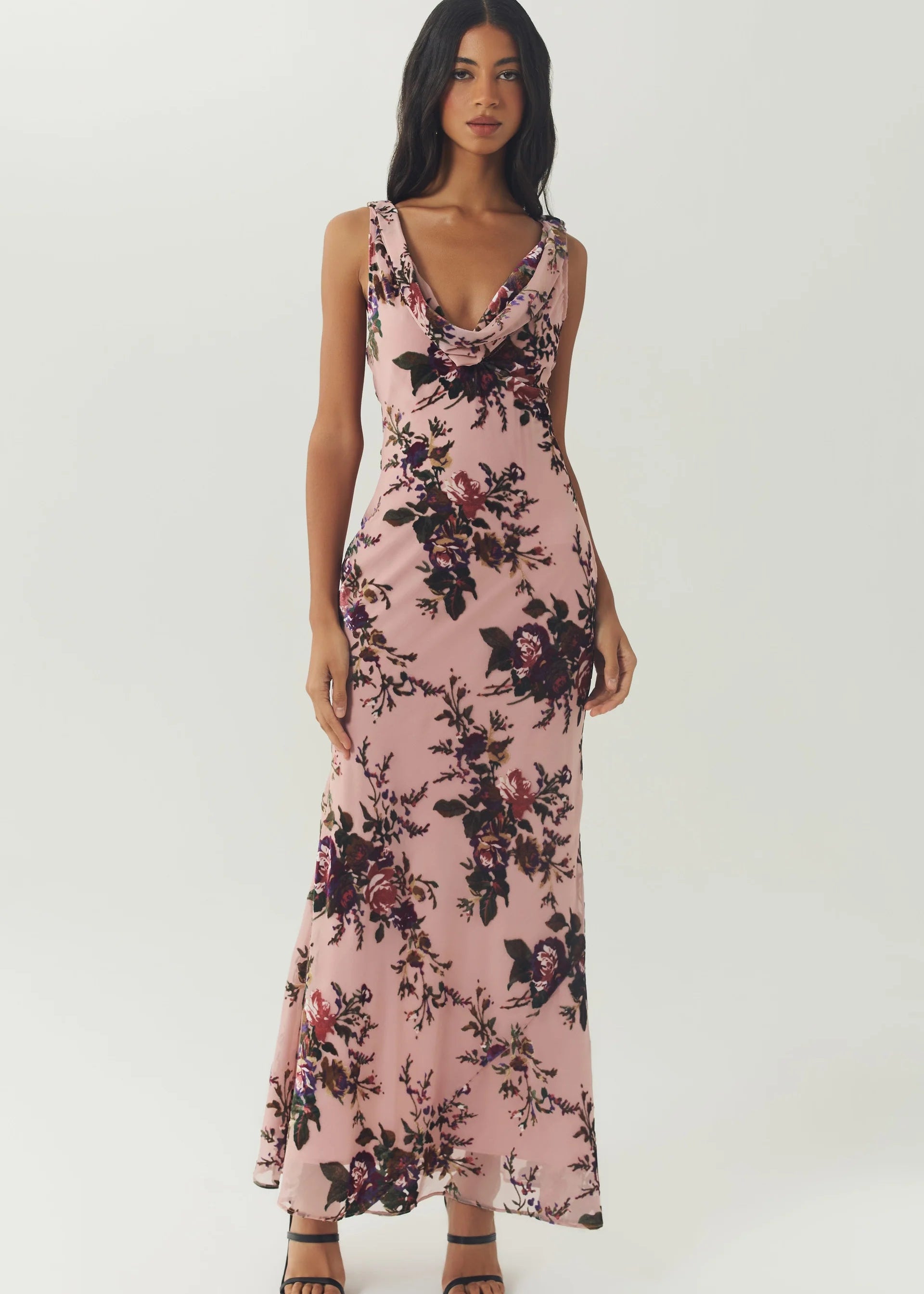 A woman modeling a sleeveless, floor-length pink maxi dress with a dark floral print, photographed against a plain light gray studio background.