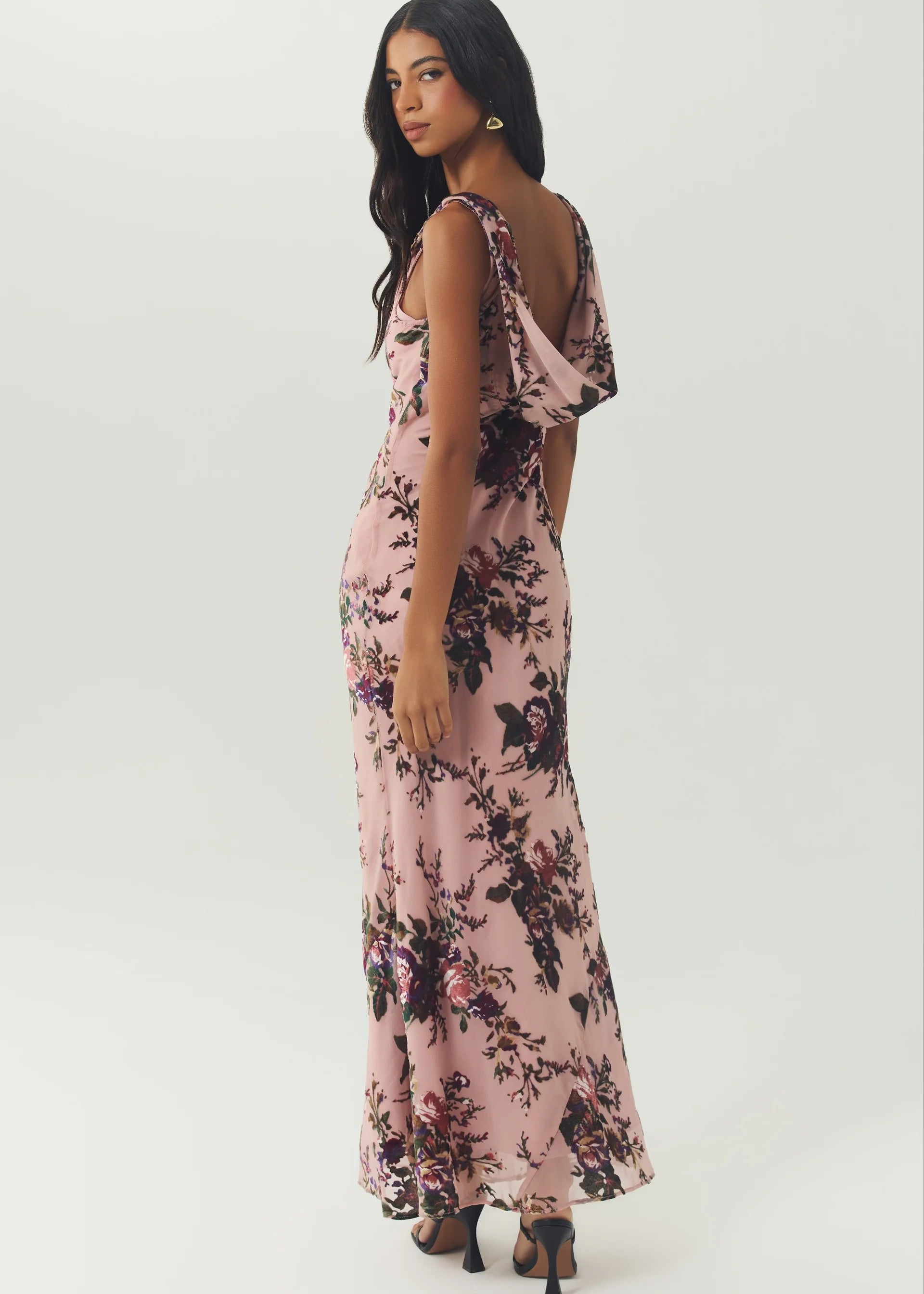 A woman modeling a sleeveless pink maxi dress with a dark floral print, shown from the back against a plain light gray studio background.