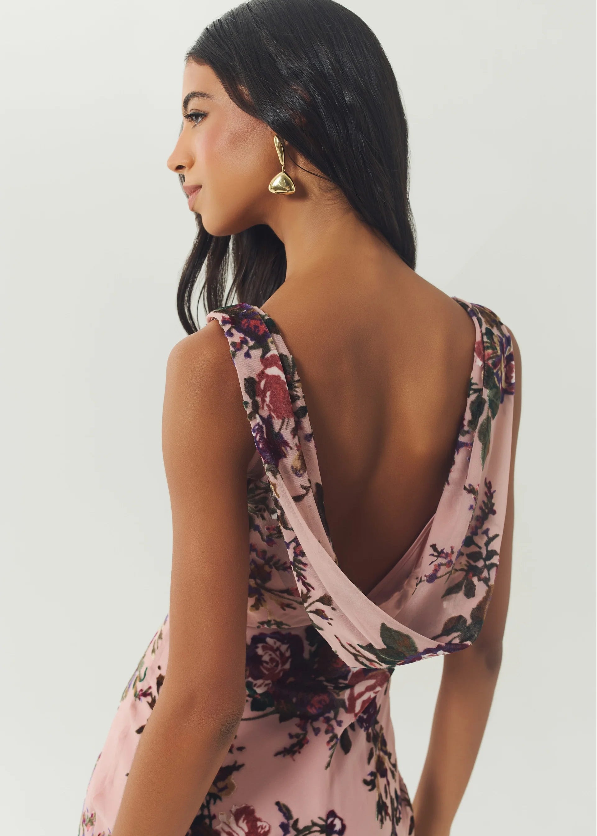 A woman shown from the back wearing a light pink floral dress with a draped open back and gold earrings, photographed against a plain light gray studio background.