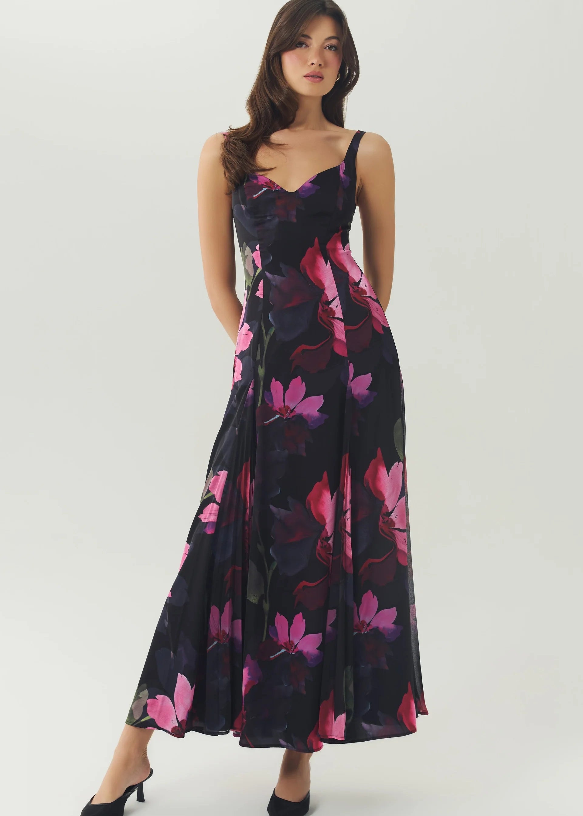 A woman modeling a sleeveless black maxi dress with a vibrant pink and purple floral print, paired with black pointed-toe heels, against a plain light gray studio background.