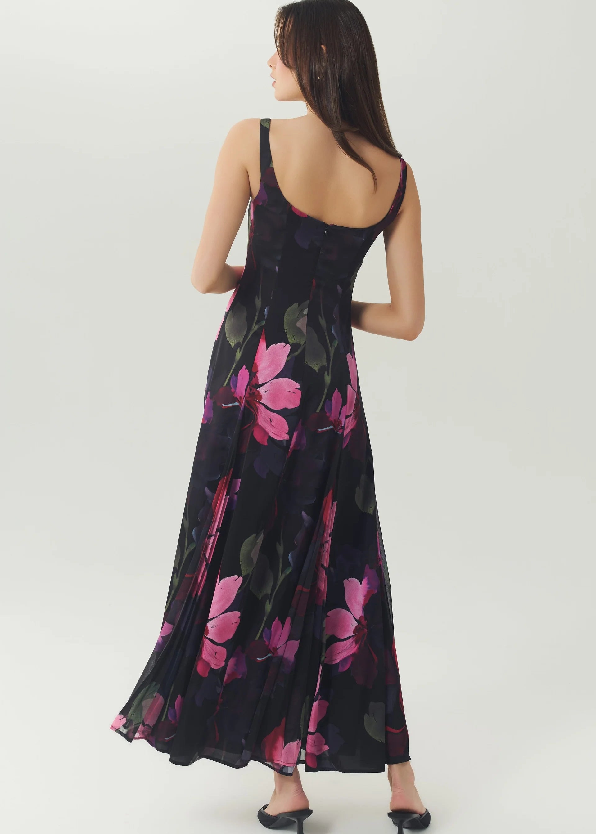 A woman wearing a sleeveless black maxi dress with a pink and green floral print, shown from the back against a light gray studio background.