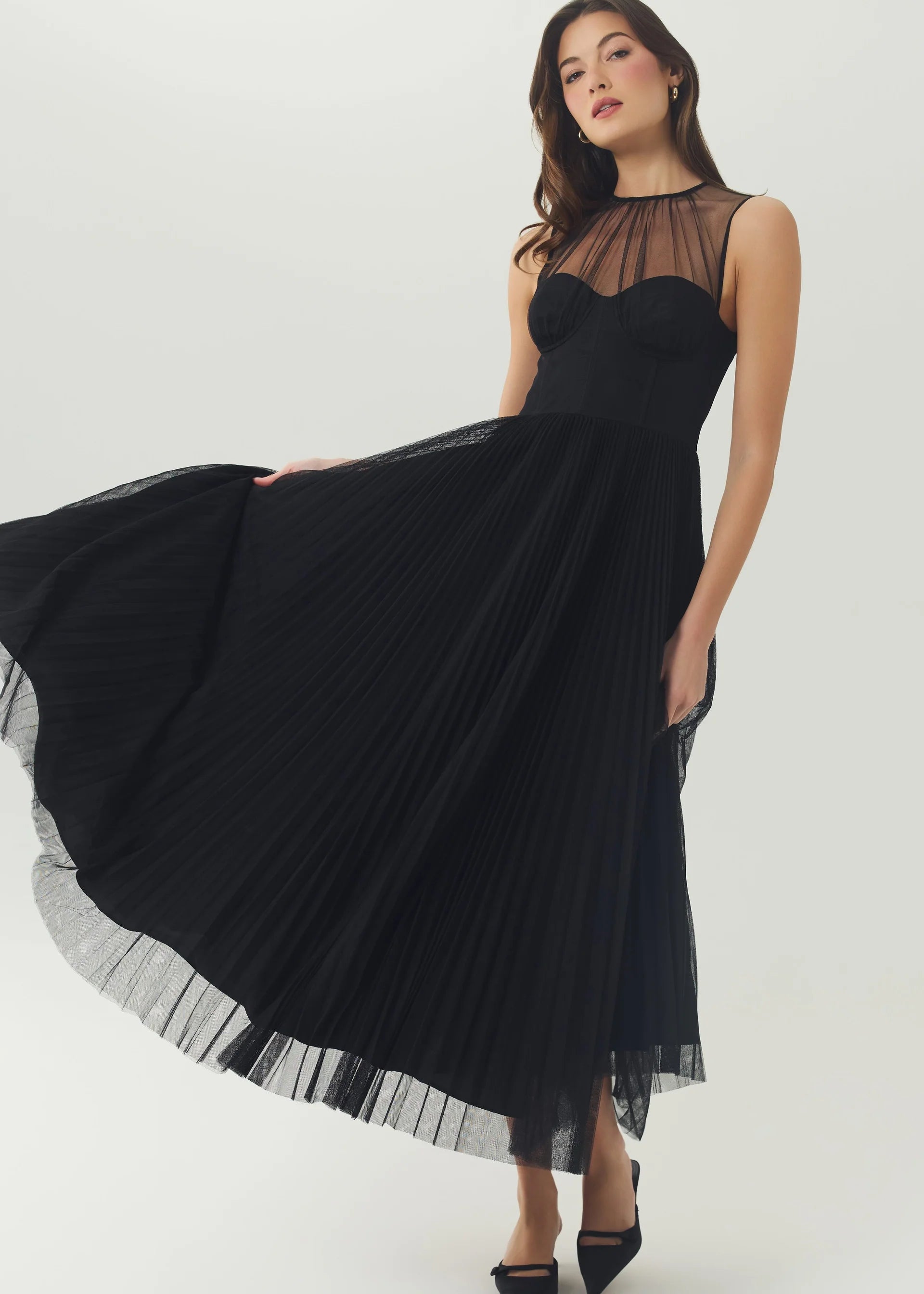 A woman modeling a sleeveless black midi dress with sheer mesh upper bodice and a flowing pleated skirt, photographed against a plain light studio background.