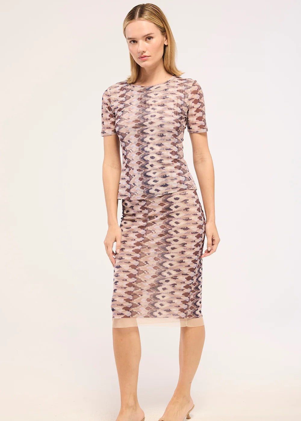 A woman modeling a short-sleeve midi dress with a beige, brown, and pink geometric pattern, paired with beige heeled sandals, against a plain light background.