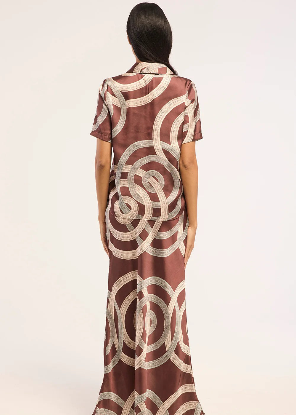 A woman viewed from behind wearing a floor-length short-sleeve dress with a brown base and cream abstract circular pattern, set against a plain light background.
