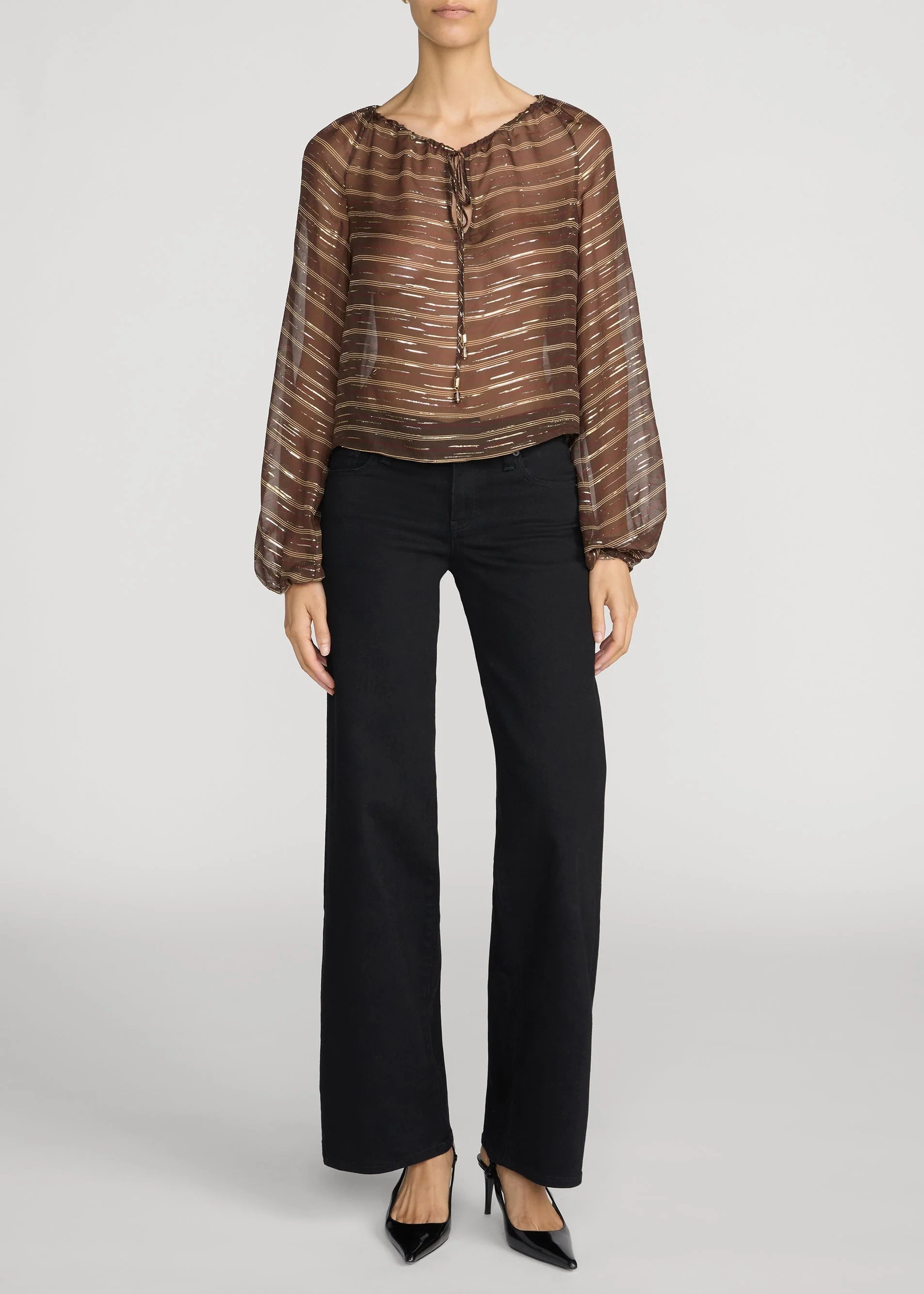 A model wearing a brown and beige striped long-sleeve blouse with a tie neckline, paired with black wide-leg trousers and black pointed-toe heels, against a light gray studio background.