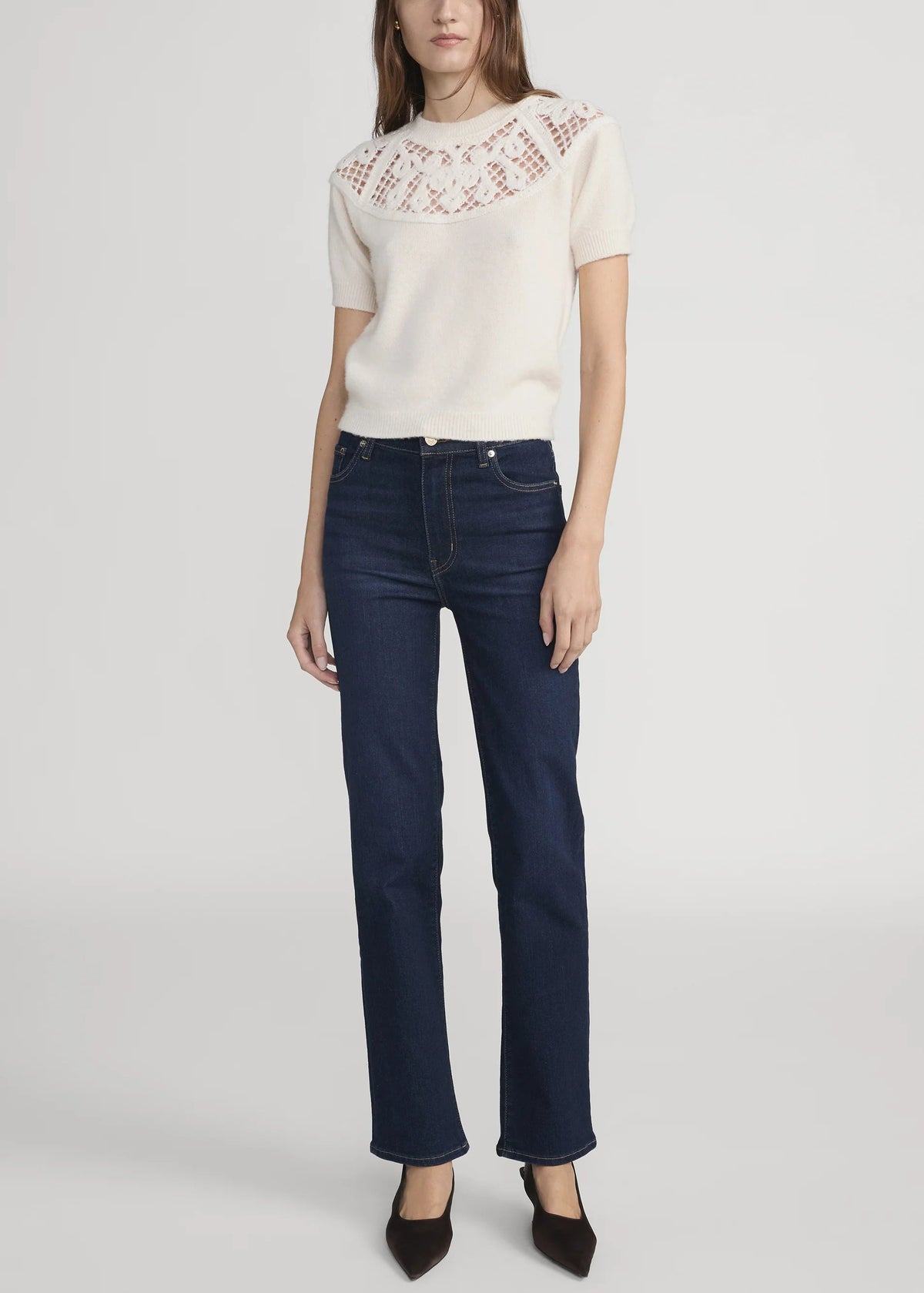 A model wearing a cream short-sleeve knit top with a textured lace-like yoke and dark blue straight-leg jeans, paired with black pointed-toe heels, against a light gray studio background.