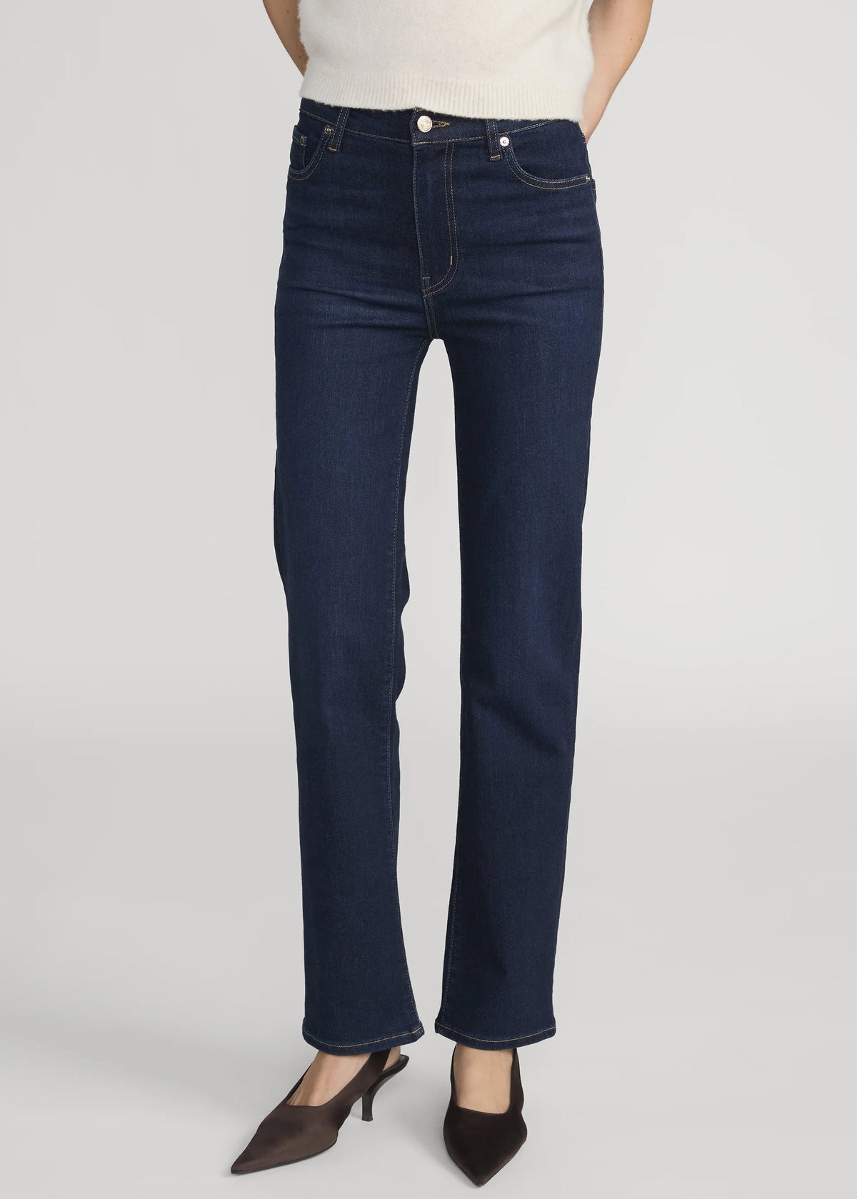 A model wearing a pair of dark blue straight-leg jeans, styled with a cream knit top and dark brown pointed-toe heels, photographed against a neutral light gray studio background.