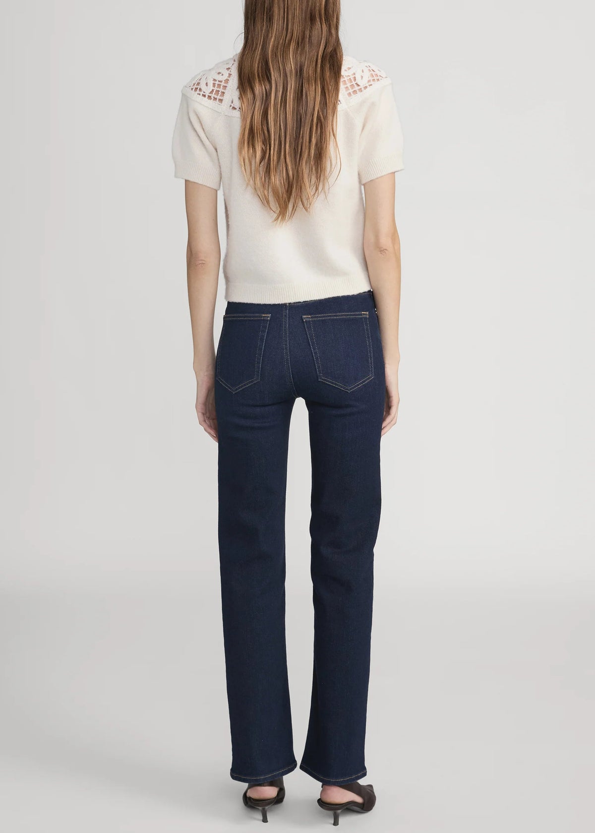 A woman viewed from the back wearing dark blue straight-leg jeans, a cream short-sleeve top with lace shoulder detailing, and black heels, set against a plain light gray studio background.