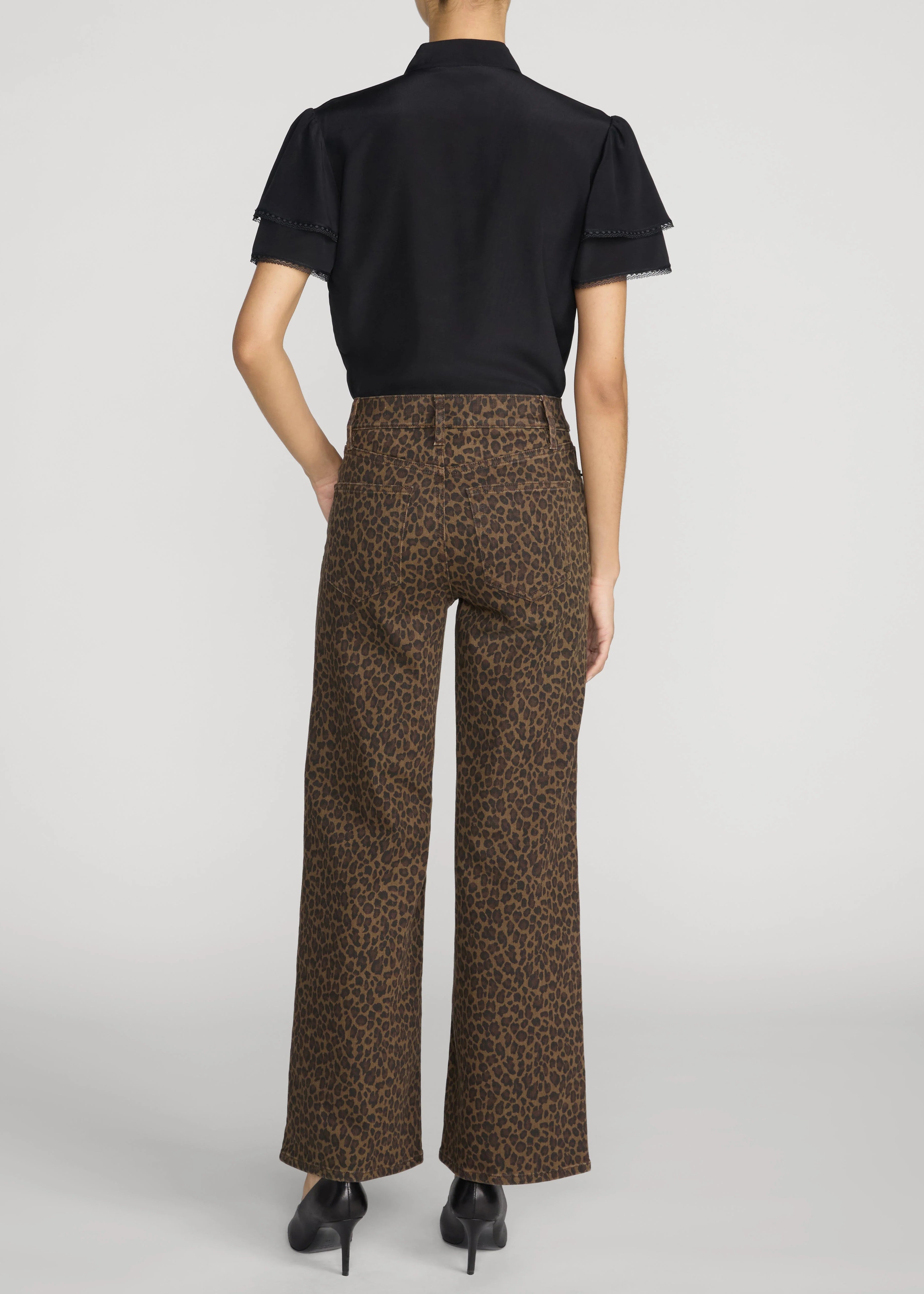 A model shown from the back wearing brown leopard-print wide-leg trousers and a black short-sleeve blouse, set against a plain light gray studio background.