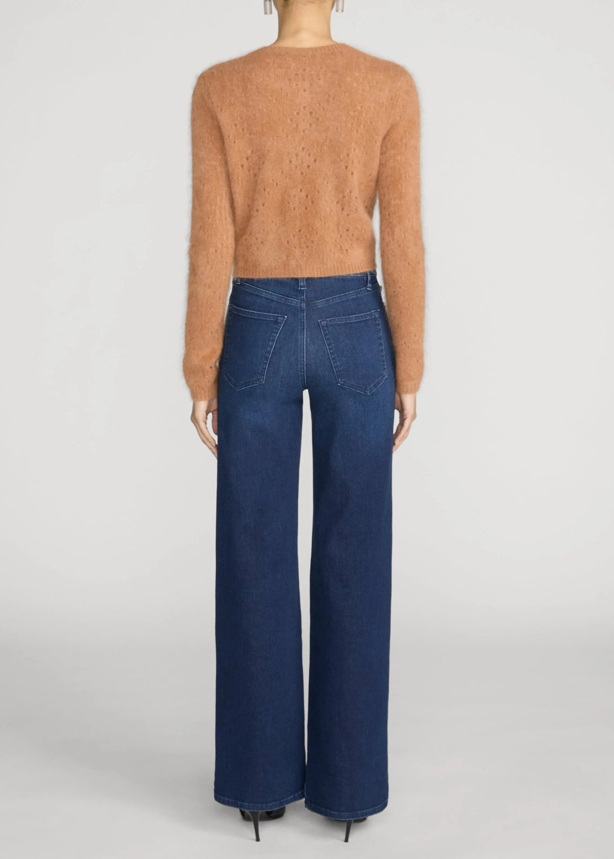 A model shown from the back wearing a fuzzy cropped knit sweater in a warm camel tan and wide-leg dark blue jeans, set against a plain light gray studio background.