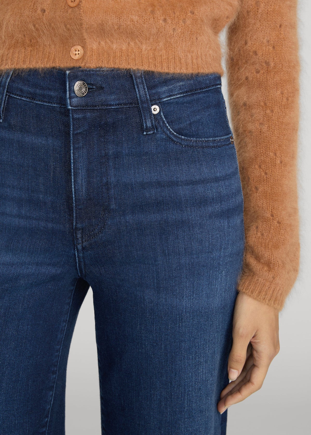 Close-up of a person wearing dark blue denim jeans and a fuzzy tan cardigan, showing the waistband, front pocket, button, and hand against a light gray background.
