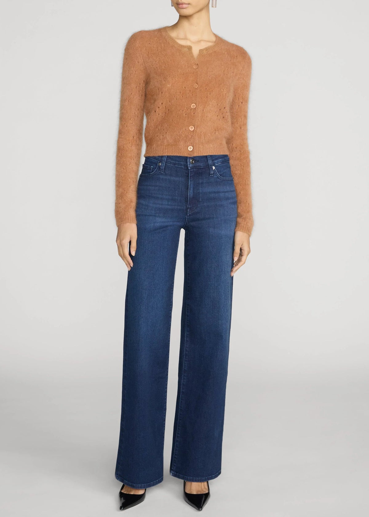 A model wearing a cropped, fuzzy tan button-up cardigan and high-waisted, wide-leg blue jeans, paired with black pointed-toe heels, against a light gray studio background.
