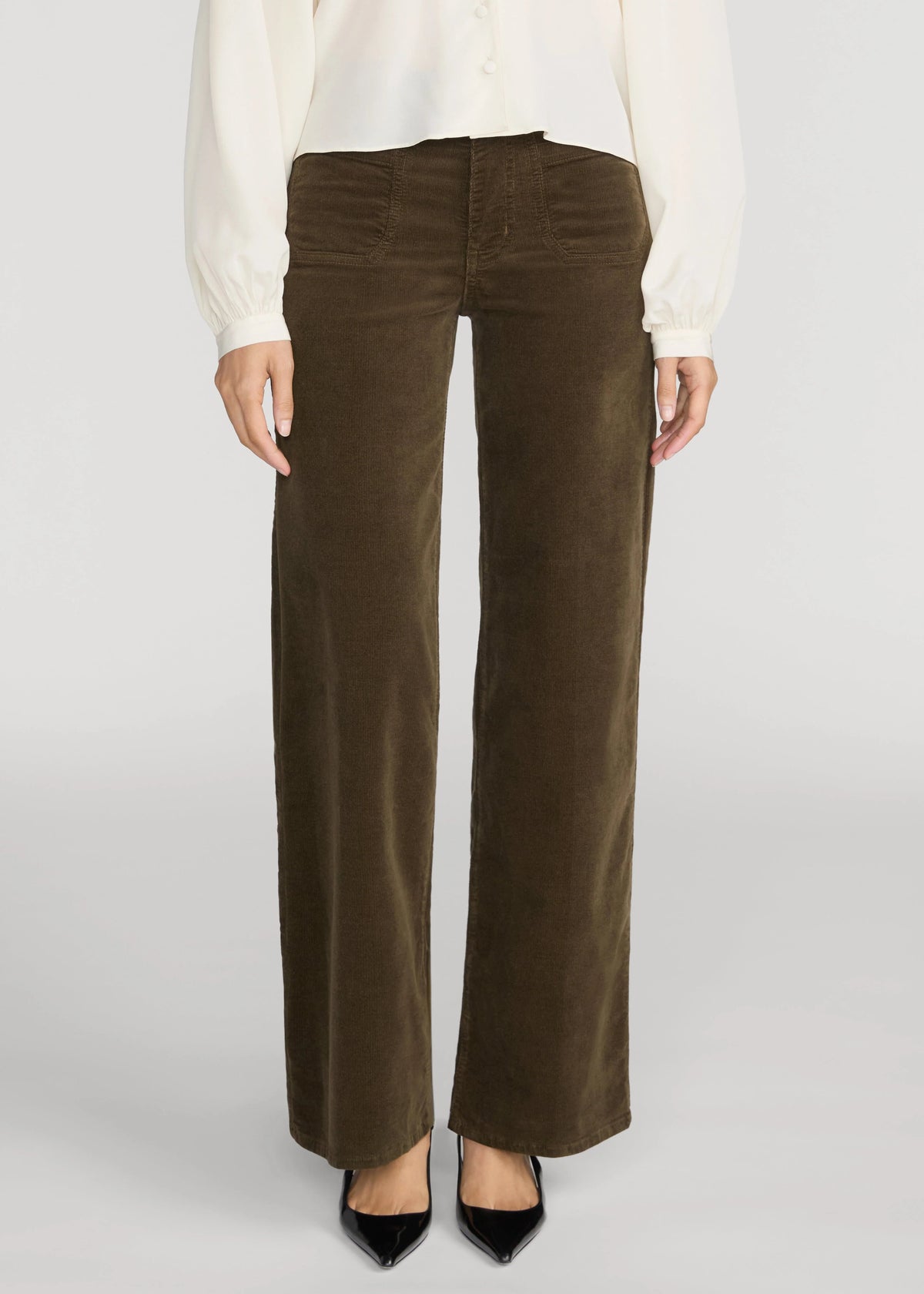 A person wearing dark brown wide-leg corduroy pants with a white blouse and black pointed shoes, shown from the waist down against a light gray studio background.