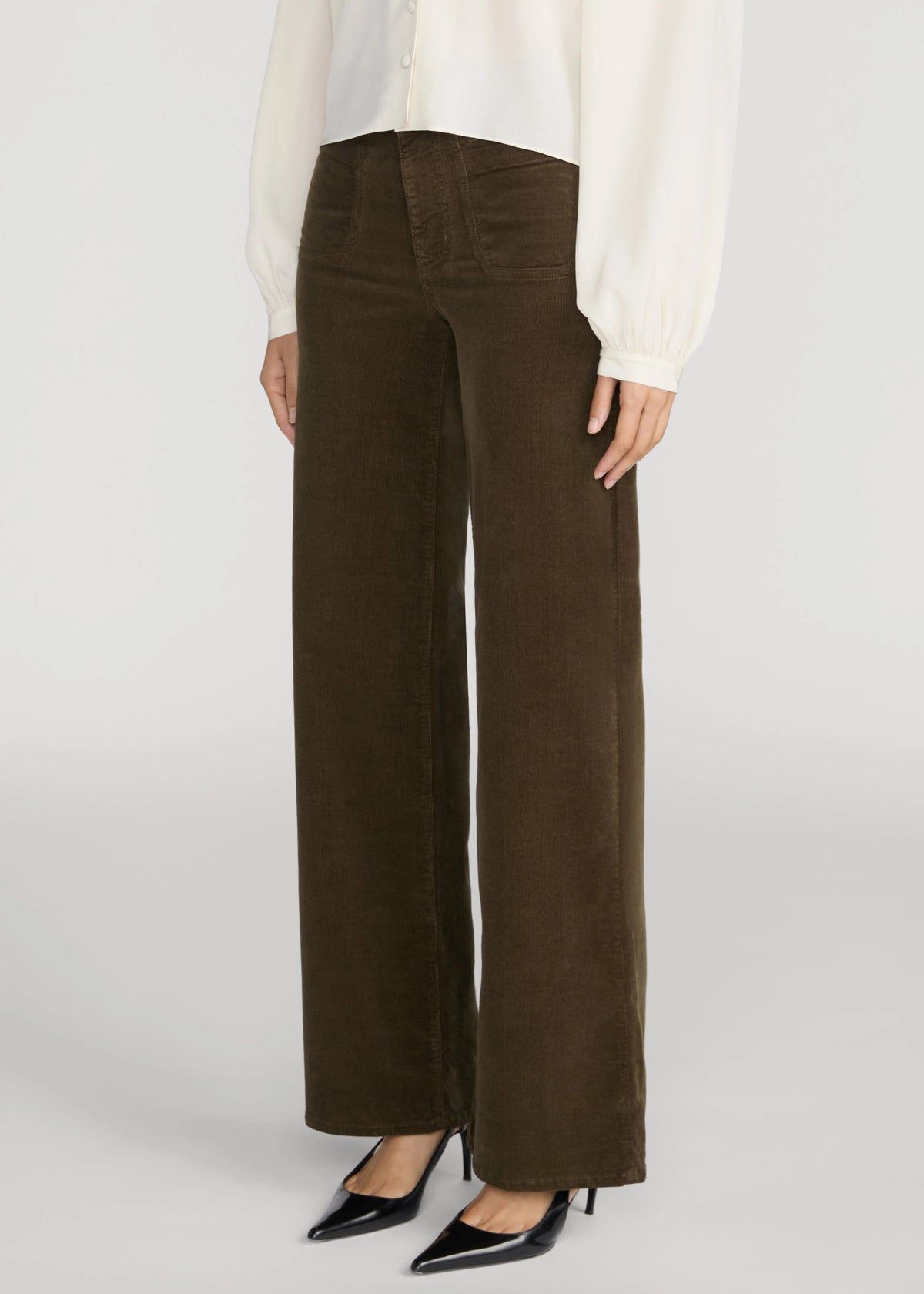 A model wearing dark brown wide-leg corduroy trousers, styled with a white blouse and black pointed-toe heels against a light gray studio background.