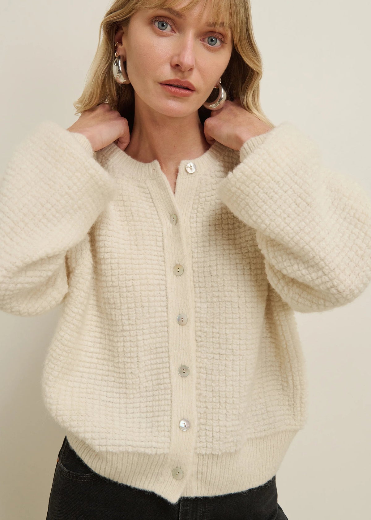 A woman wearing a cream-colored, textured waffle-knit cardigan sweater with a button-up front, paired with black pants, against a plain beige background.