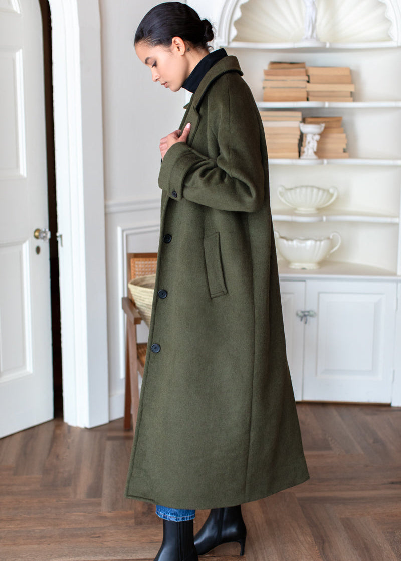 A woman wearing a long olive green wool coat over a black turtleneck, styled with blue jeans and black ankle boots, standing in a bright room with wooden floors and white shelves.