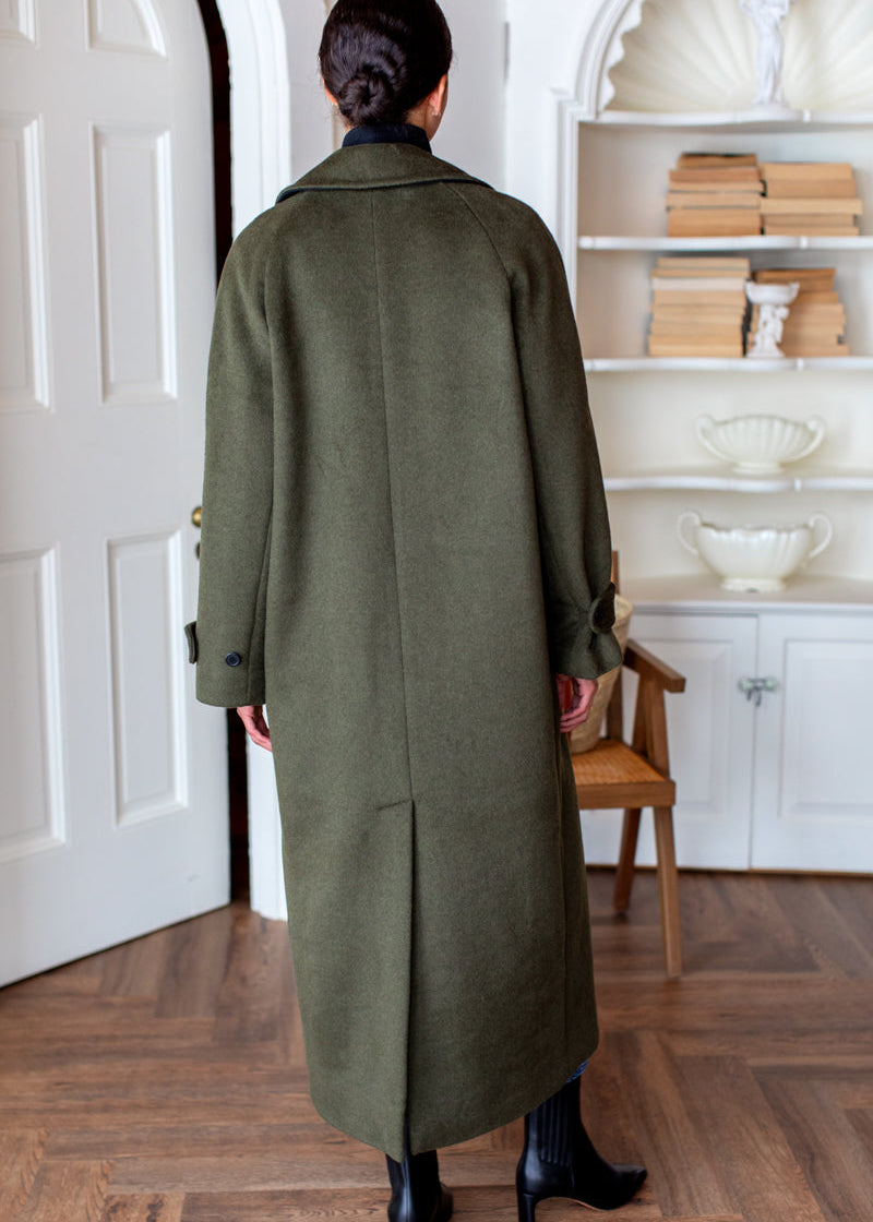 A person viewed from behind wearing a long olive green wool coat in an indoor room with wood floors, a chair, and white shelving.