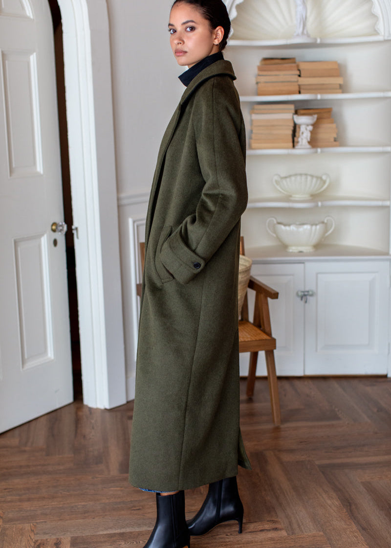 A woman stands in a bright room wearing a long olive green coat over a dark turtleneck, paired with black pointed ankle boots. The room features a wooden herringbone floor, white walls, and built-in shelves with books and decorative items.