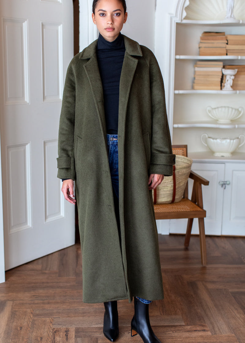 A woman stands indoors wearing a long olive green coat over a black turtleneck, blue jeans, and black ankle boots. The room has wood floors, a white door, and a white shelving unit in the background.