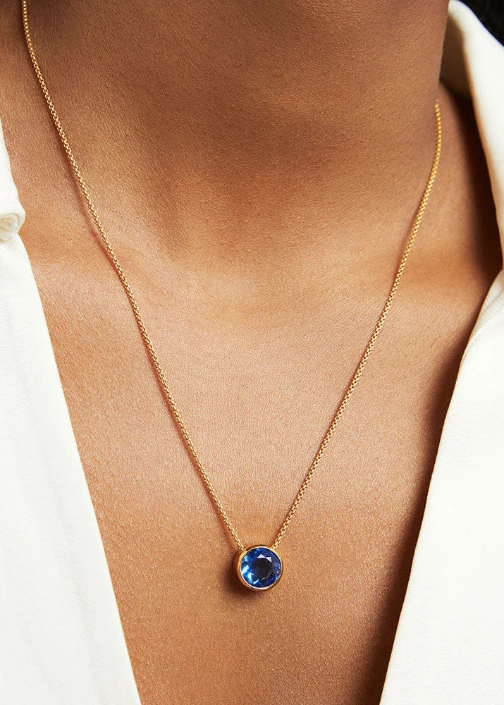 A close-up of a person wearing a delicate gold chain necklace with a round blue gemstone pendant, styled with a white collared shirt.