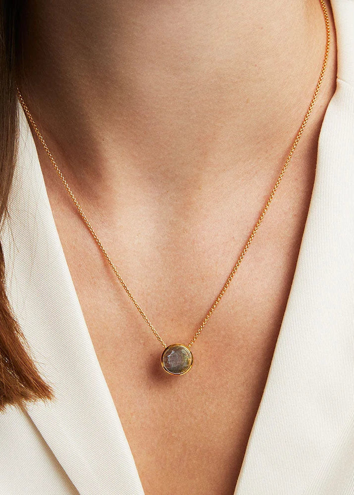 A close-up of a model wearing a delicate gold chain necklace with a round, iridescent gray pendant, styled with a white blazer.