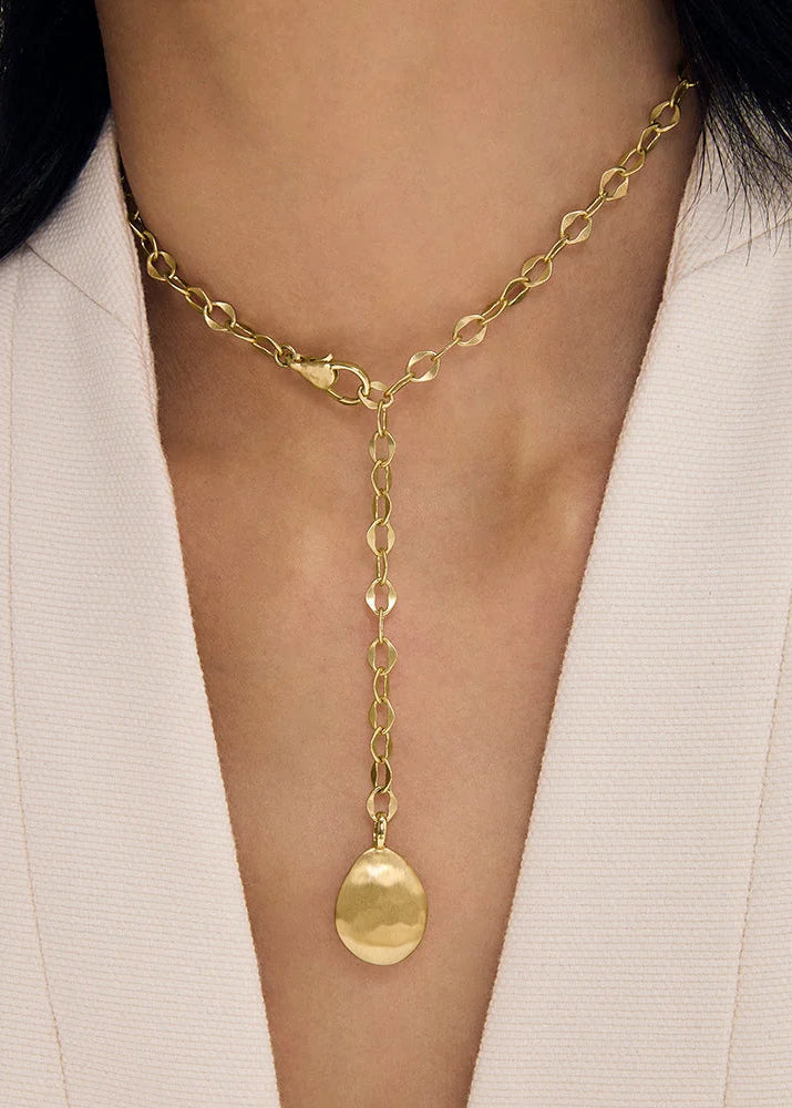 A close-up of a model wearing a gold Y-necklace with a textured hammered teardrop pendant, styled with a white blazer.